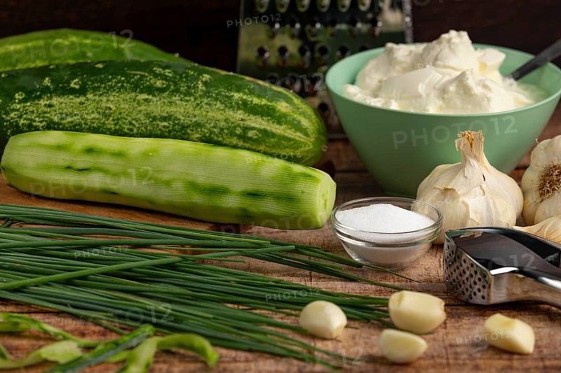 Fresh ingredients such as peeled cucumbers, garlic, chives and sour cream on a wooden table with kitchen utensils