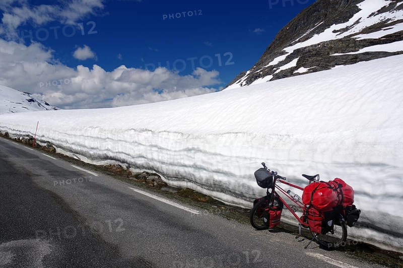 A bicycle with red panniers stands next to a snow-covered mountain road under a clear sky, Fjell in spring, Geiranger, Langevatn, Norway, Europe