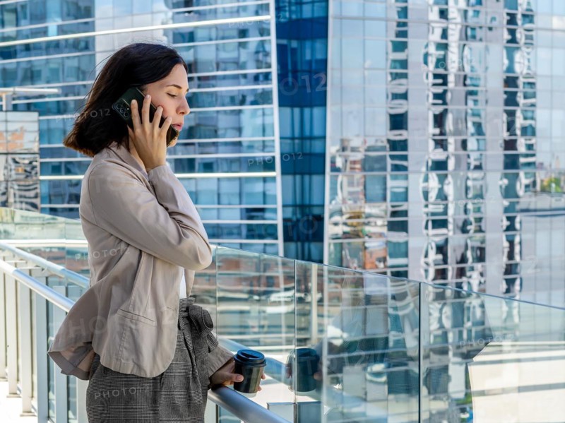 A woman is standing on a balcony, holding a paper cup of coffee and talking on her cell phone during her office break