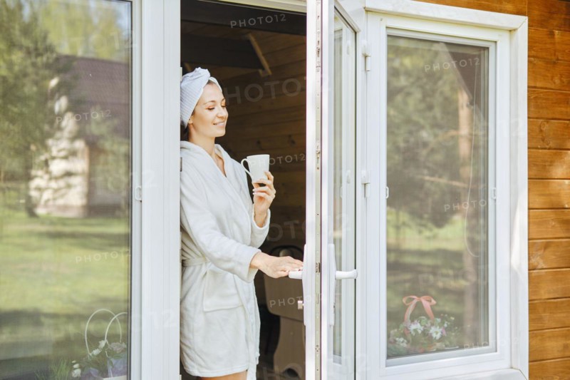 Smiling woman in bathrobe and cup of coffee in her opens the door onto terrace of wooden cabin house