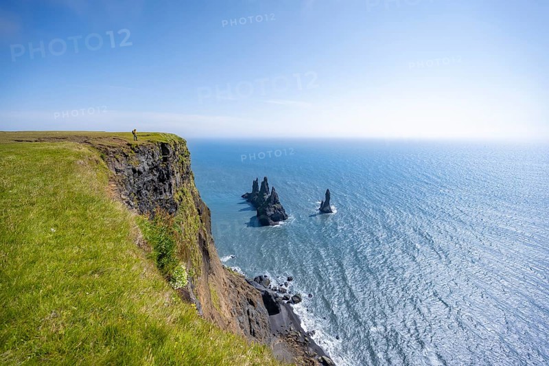 Young man standing on cliff, view from steep cliff to the rocks Reynisdrangar in the water, at Reynisfjara beach, Vik, South Iceland, Iceland, Europe