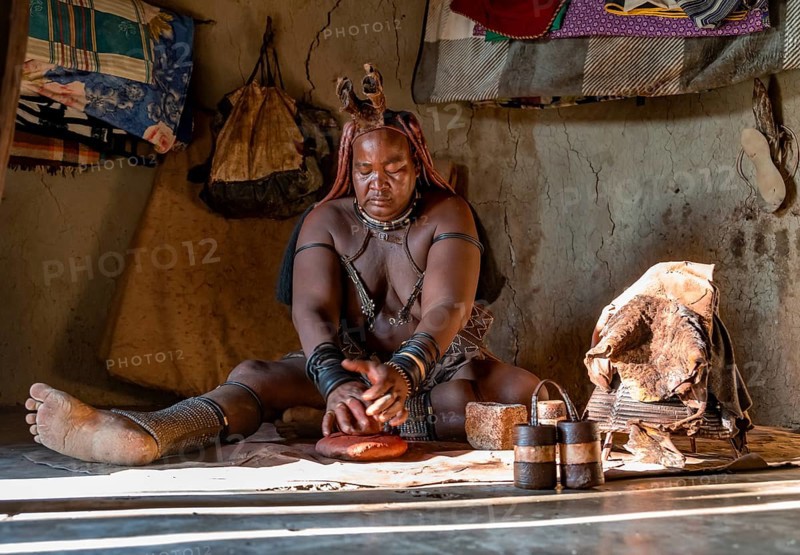 Traditional Himba woman making ochre body paint from fat and ochre, interior of the first woman's mud hut in a Himba village, traditional containers for ochre paint and woven frame with leather for body care with smoke, near Opuwo, Kaokoveld, Kunene, Namibia, Africa