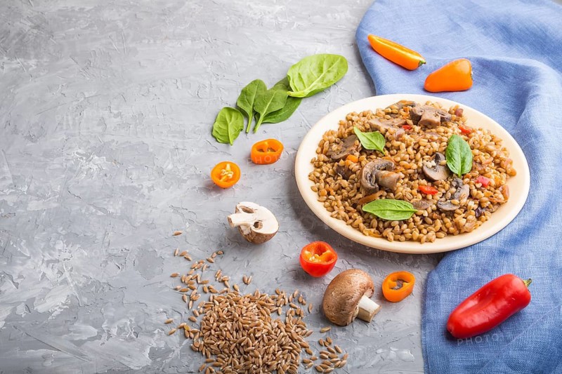 Spelt (dinkel wheat) porridge with vegetables and mushrooms on ceramic plate on a gray concrete background and blue linen textile. Side view, copy space. Russian traditional cuisine