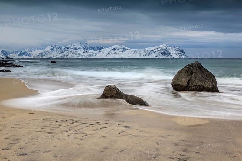Rocks on the beach in front of snowy mountains, dark clouds, sea, spray, winter, Vikten, Flakstadoya, Lofoten, Norway, Europe
