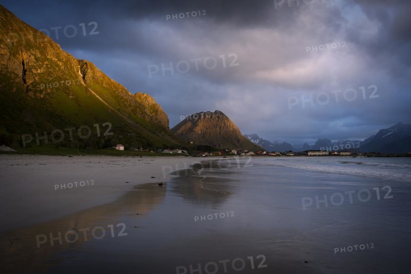 Landscape on the Lofoten Islands. The village of Ramberg, with the sea and the sandy beach (Rambergstranda) in front. Mountains in the background. Partial reflection. At night at the time of the midnight sun. Clouds in the sky. Mountains in the sunlight. Early summer. Ramberg, Flakstadoya, Lofoten, Norway, Europe