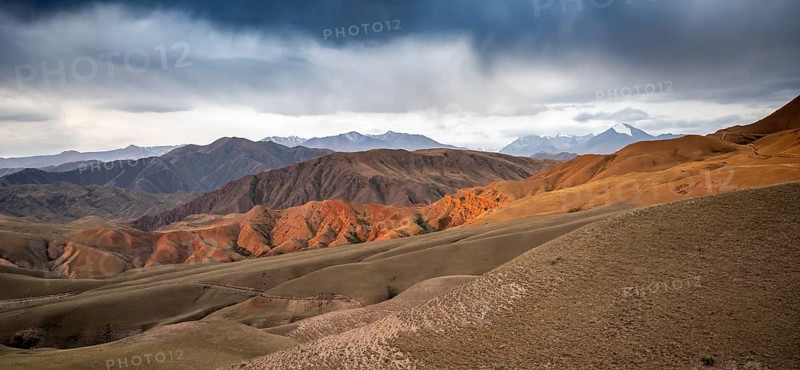 View over eroded mountainous landscape of Konorchek Canyon with red sandstone cliffs, high mountains with snow behind, dramatic dark clouds, Konorchek Canyon, Chuy, Kyrgyzstan, Asia