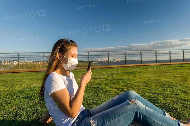 Young woman sitting on the grass with a protective mask during the SARS CoV-2 pandemic season (COVID 19), using her smartphone on the new Orla do Guaiba in Porto Alegre on a beautiful sunset day