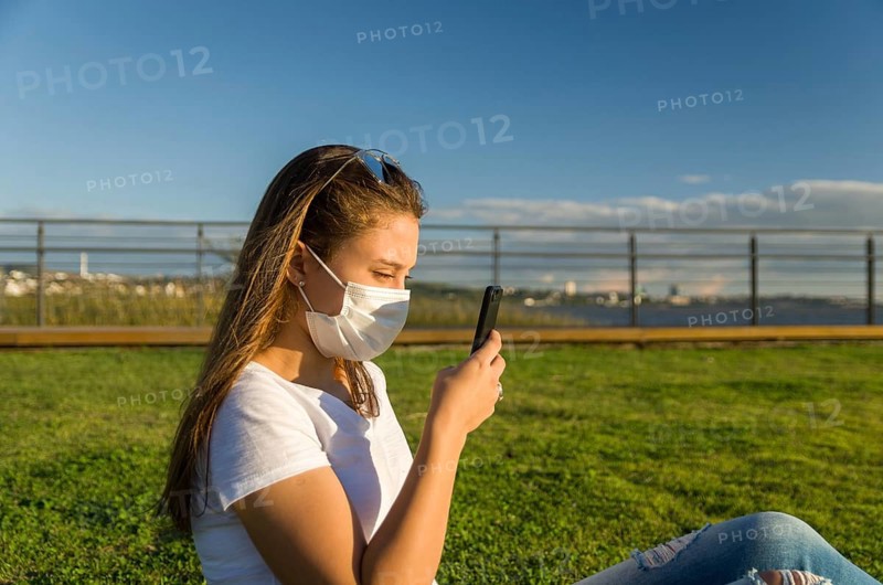 Young woman sitting on the grass with a protective mask during the SARS CoV-2 pandemic season (COVID 19), using her smartphone on the new Orla do Guaiba in Porto Alegre on a beautiful sunset day