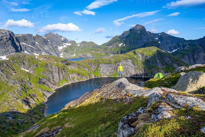 Mountaineer standing on a rock with green camping tent, mountain landscape with lake Tennesvatnet, at sunrise, behind peak of Hermannsdalstinden, Moskenesoya, Lofoten, Nordland, Norway, Europe