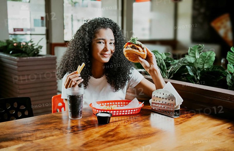 Happy girl enjoying a delicious hamburger sitting in a restaurant. Portrait of young afro woman holding a hamburger with fries in a restaurant