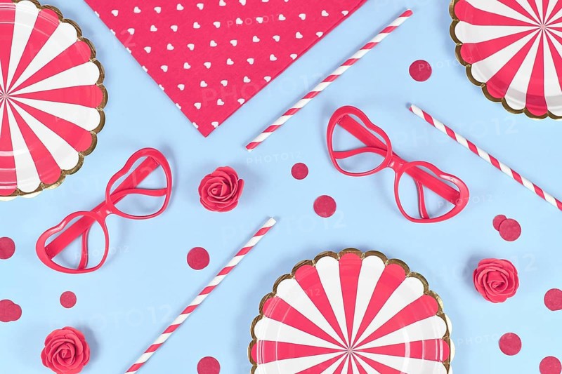Party flat lay with paper plates, heart shaped glasses, roses, confetti and drinking straws on blue background