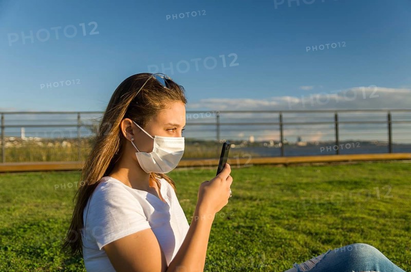 Young woman sitting on the grass with a protective mask during the SARS CoV-2 pandemic season (COVID 19), using her smartphone on the new Orla do Guaiba in Porto Alegre on a beautiful sunset day