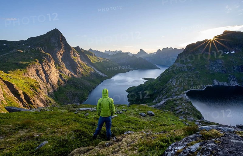 Mountaineer looking over mountain landscape with pointed mountain peaks and fjord Forsfjorden with village Vindstad, sun star at sunrise, Moskenesoya, Lofoten, Nordland, Norway, Europe