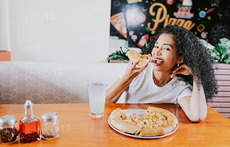 Smiling young woman enjoying a pizza in a restaurant. Happy afro hair girl eating pizza in a restaurant, eating and looking at camera