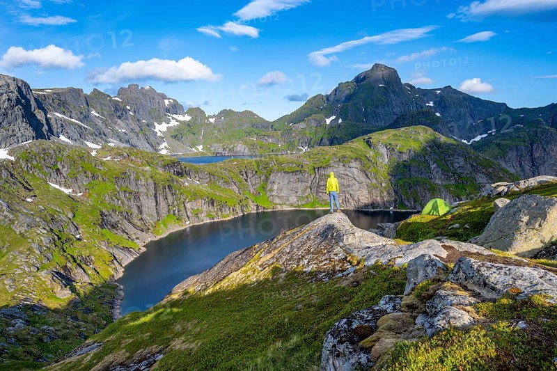 Mountaineer standing on a rock with green camping tent, mountain landscape with lake Tennesvatnet, at sunrise, behind peak of Hermannsdalstinden, Moskenesoya, Lofoten, Nordland, Norway, Europe