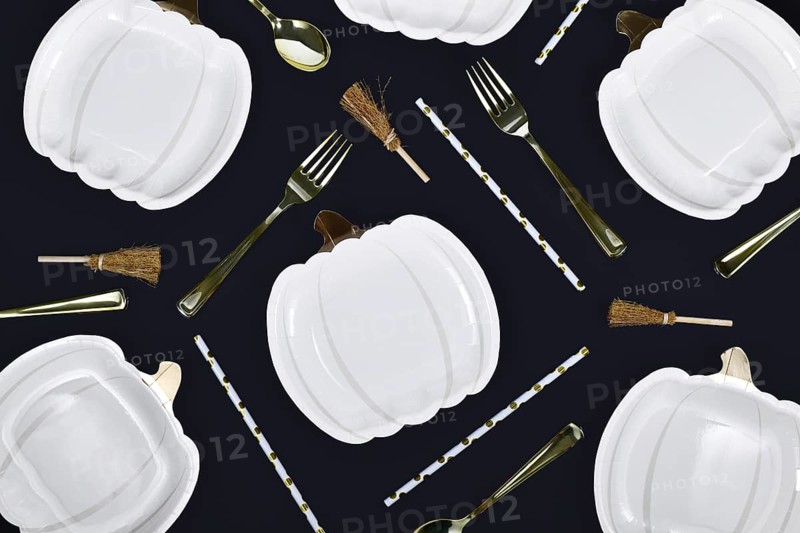 Elegant Halloween party flat lay with pumpkin shaped plates, brooms and golden forks and spoons on black background