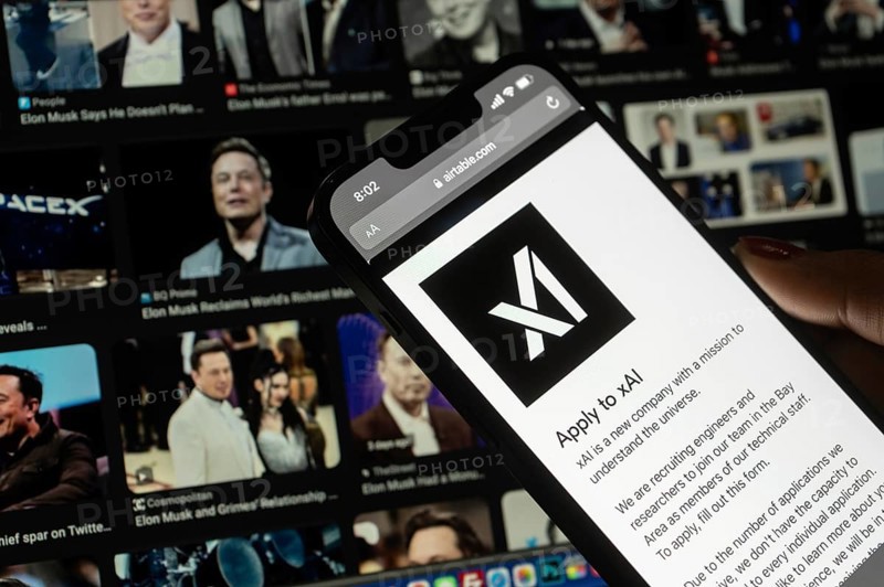 In this photo illustration, A person browses website of xAI, displayed on a mobile phone, Elon Musk images in the background screen, in Guwahati, India 0n 15 July 2023. Tesla CEO Elon Musk launches Artificial Intelligence company xAI on 12 July 2023, the company's goal is to understand the true nature of the universe
