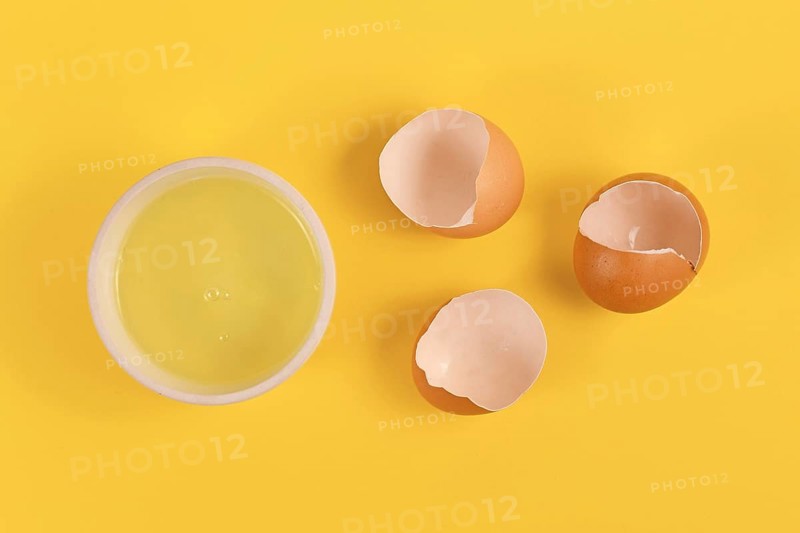 Bowl with egg white next to broken eggshells on yellow background