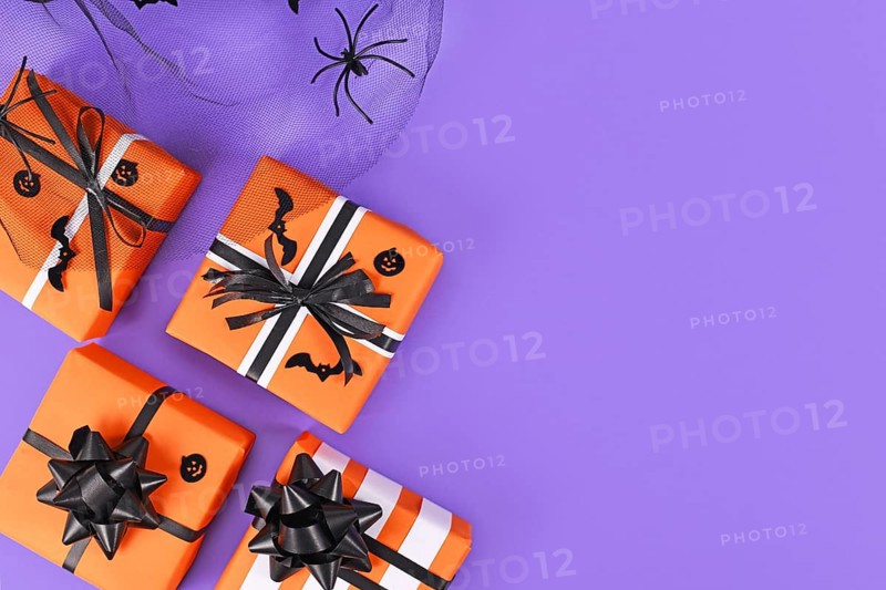 Gift boxes with traditional Halloween event colors orange, black and white and small bats and pumpkins and spider on purple background with empty copy space