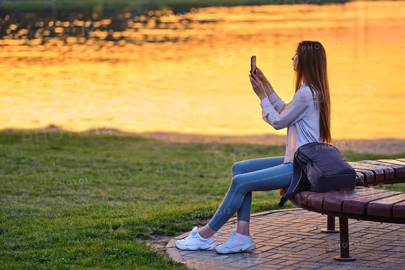 Blogger woman streaming online the beautiful sunset sitting on bench by river