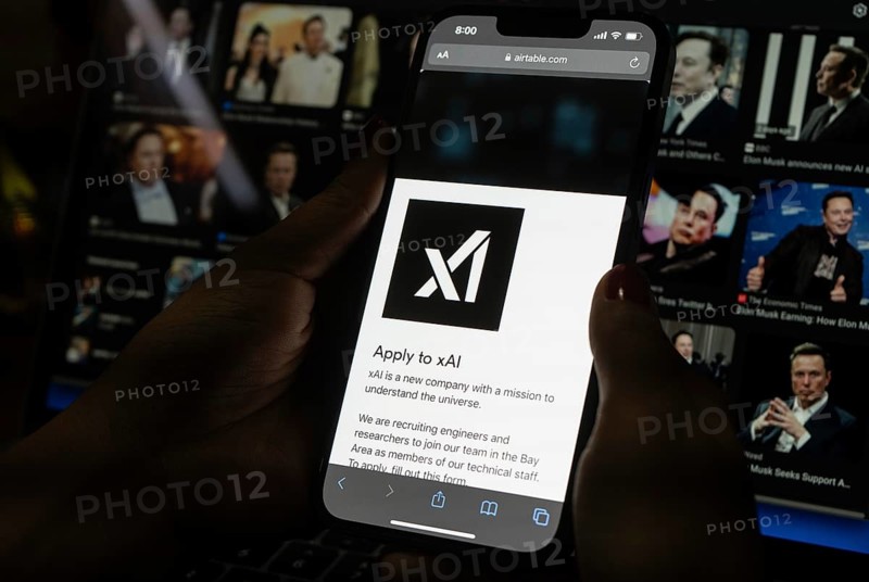 In this photo illustration, A person browses website of xAI, displayed on a mobile phone, Elon Musk images in the background screen, in Guwahati, India 0n 15 July 2023. Tesla CEO Elon Musk launches Artificial Intelligence company xAI on 12 July 2023, the company's goal is to understand the true nature of the universe