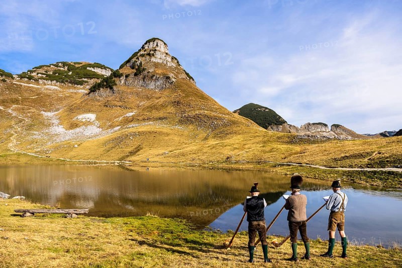 Three men play the alphorn at Lake Augstsee on Mount Loser. The Austrian alphorn trio Klangholz. Mount Atterkogel in the background. Autumn, good weather, blue sky. Reflection. Altaussee, Bad Aussee, Ausseer Land, Totes Gebirge, Styria, Upper Austria, Austria, Europe