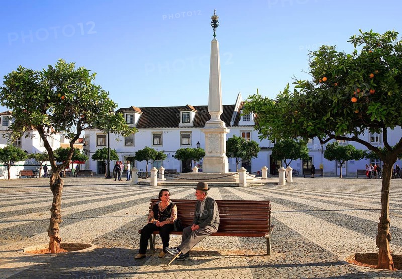 Praca do Marques de Pombal square in Vila Real de Santo Antonio, Algarve, Portugal, Elderly people sitting on a bench in a sunny market square with an obelisk and white buildings in the background, Europe