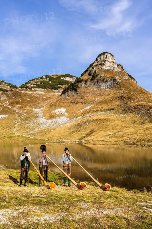 Three men play the alphorn at Lake Augstsee on Mount Loser. The Austrian alphorn trio Klangholz. Mount Atterkogel in the background. Autumn, good weather, blue sky. Reflection. Altaussee, Bad Aussee, Ausseer Land, Totes Gebirge, Styria, Upper Austria, Austria, Europe