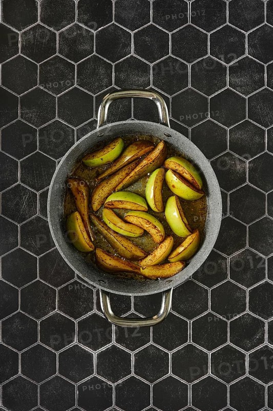 Sliced apples and pears caramelizing in a skillet with melted duck fat glaze on a hexagonal tile background