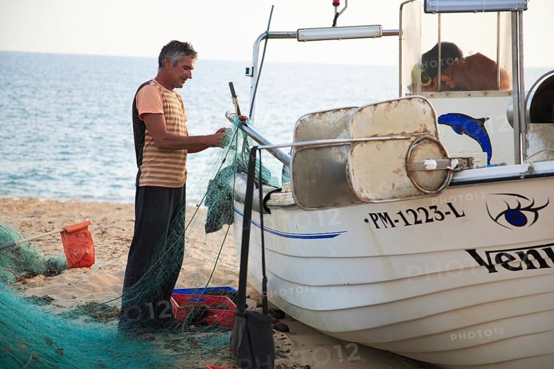 Fisherman in Armacao de Pera, Algarve, Portugal, fisherman preparing a net next to a boat on the beach. In the background the sea and bright sunlight, Europe