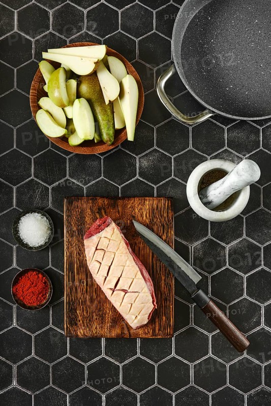 Overhead view of raw duck breast with the skin cut crosswise on wooden chopping board surrounded with apple and pear slices in a bowl