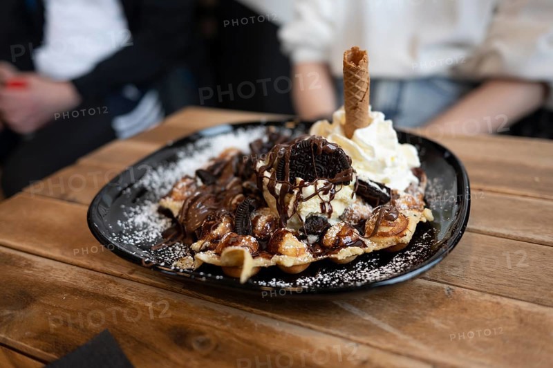 Belgian waffle with vanilla ice cream and whipped cream, waffle rolls, chocolate sauce and Oreo biscuit served on a black plate and dusted with icing sugar, on a wooden table, slight top view, male and female person recognisable in the background, Wallonia, Belgium, Europe