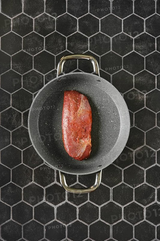Top view of a raw duck breast lying on a non-stick pan with two handles, against a background of black tiles