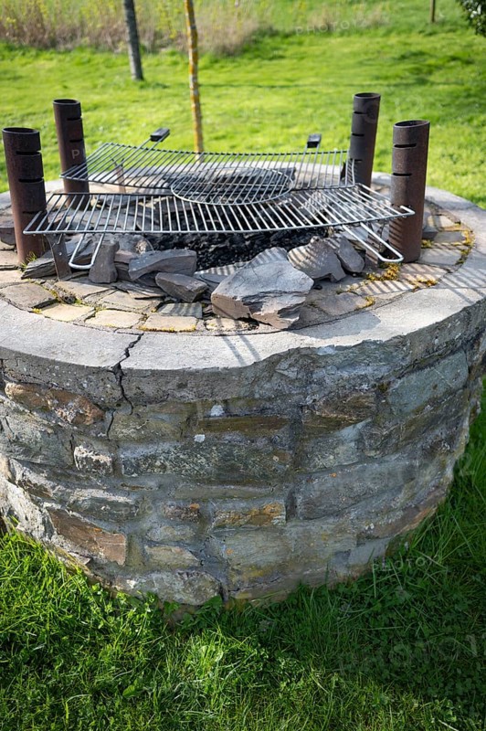 Outdoor barbecue, barbecue from an old well, two used grill grates lying on top, four metal pipes outside, burnt coal visible, stones, green meadow in the background and foreground, Wallonia, Belgium, Europe