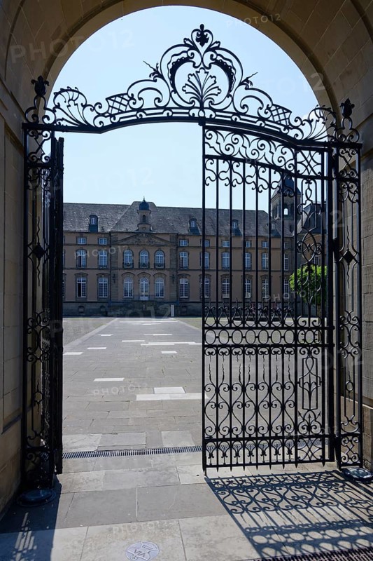 Iron gate opens onto a sunny courtyard with historic building, Echternach Abbey, Echternach, Luxembourg, Europe