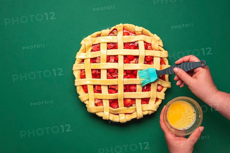 Woman hands washing with egg yolk the pie crust. Top view with strawberries and rhubarb tart. Uncooked berries pie