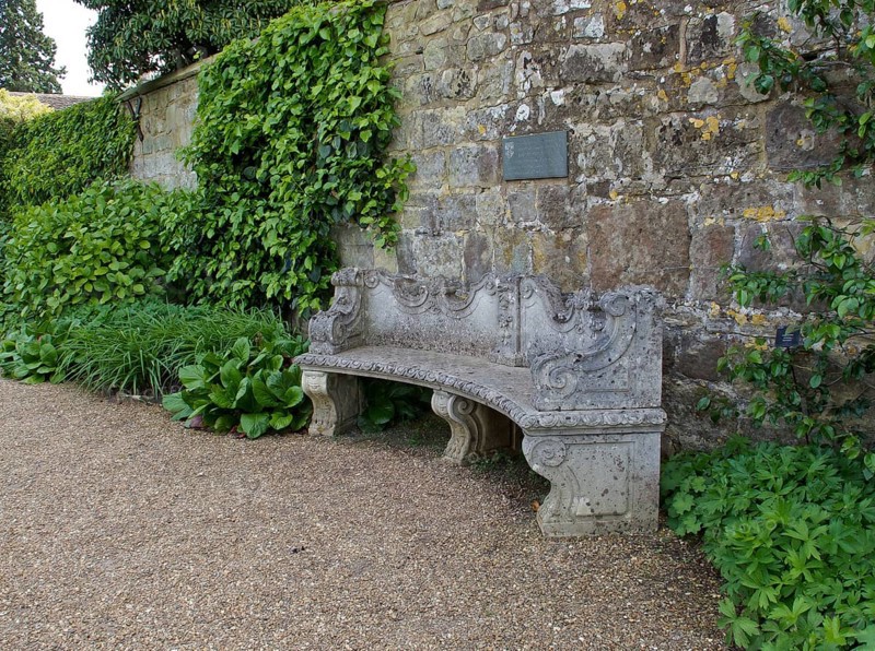 Old curved and ornately designed stone garden seat against old stone wall with green foliage