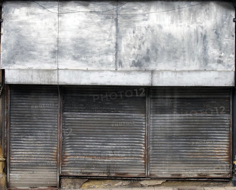 The front of an abandoned store on a street with closed dirty rusting metal shutters over the shop front and door