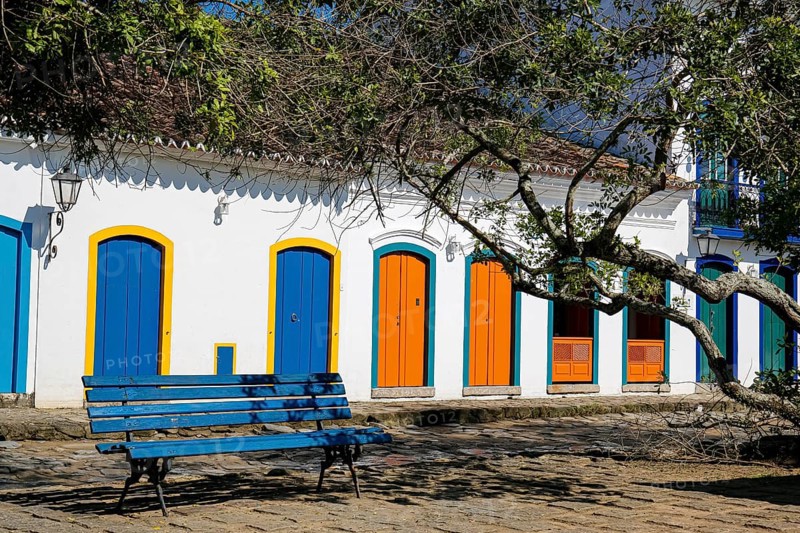 View to colonial house facades with colorful wooden doors in sunshine with a blue bench and tree branches in front, historic town Paraty, Brazil, Unesco World Heritage, South America