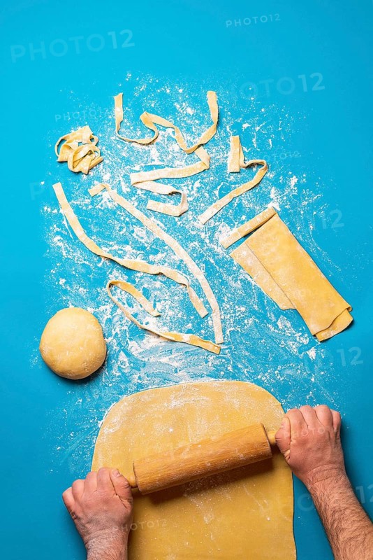 Man rolling semolina dough to make pasta, on a blue background. Top view of making tagliatelle pasta. Authentic Italian food recipe. Preparing dinner