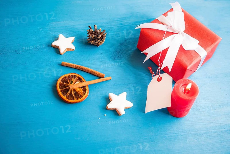 Gift, wrapped in red paper and tied with white ribbon and bow, with a blank tag attached to it, surrounded by cinnamon sticks, dried orange and star shaped cookies