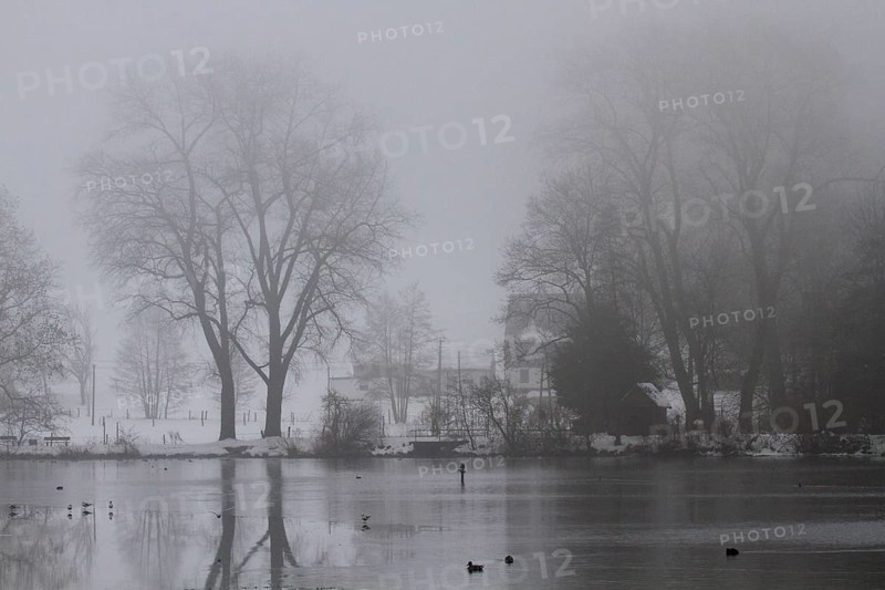 In foggy weather a lake with a house and trees in the background
