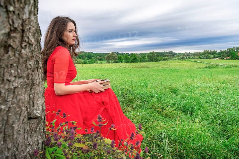 Beautiful woman, in an elegant red dress, sitting on a bench under a tree, with a book and pocket watch in her hands, in Schwabisch Hall, Germany, Europe