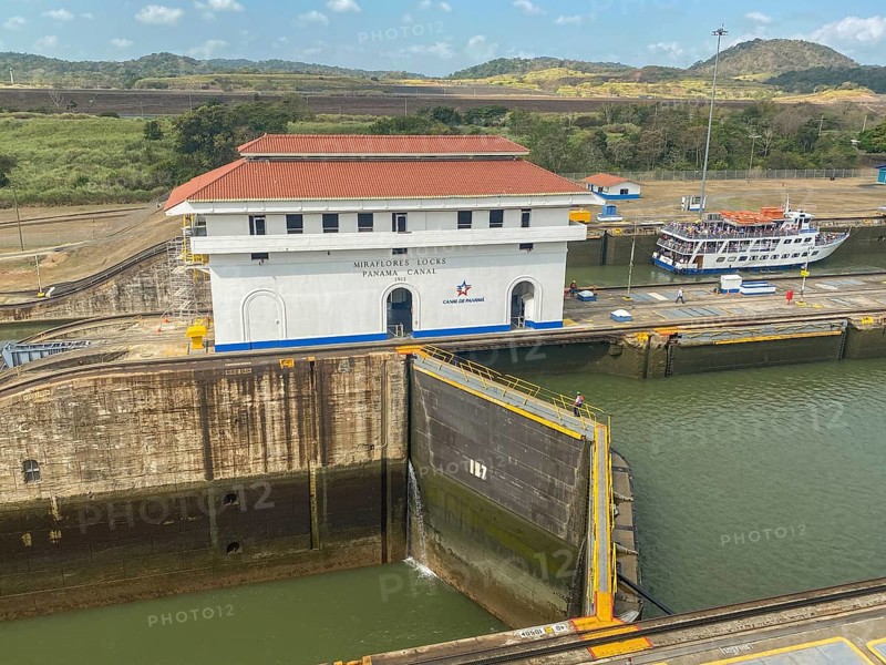 A picture of a lock with a large white building and a small boat. The surroundings are green and hilly, panama city, panamakanl, panama, Central America