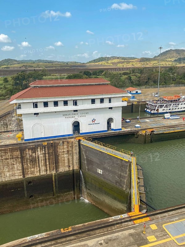 A canal lock next to a large white building. The surroundings consist of green hills and trees under a clear sky, panama city, panamakanl, panama, Central America