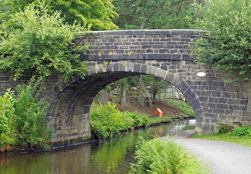 An old stone bridge crossing the rochdale canal at mytholmroyd west yorkshire with a waterside pathway surrounded by trees and bushes