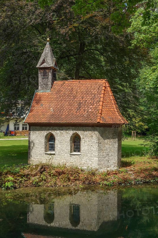 A small stone house with a red tiled roof stands on the edge of a pond, surrounded by dense forest, Alps, Austria, Europe