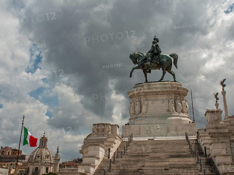 An imposing equestrian statue on a pedestal, flanked by an Italian flag and steps under a cloudy sky, rome, italy