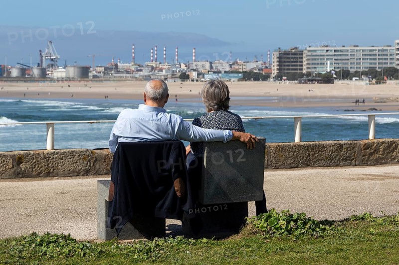 A couple of senior citizens sitting on a park bench looking at the surf in the Atlantic Ocean on the seafront at Praia de Matosinhos beach in Matosinhos, Norte region, Porto district, Portugal, Europe
