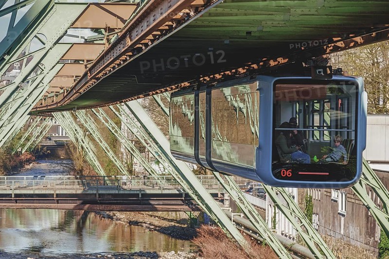 A suspension railway crosses a river in a city, surrounded by architectural metal structures in daylight, suspension railway at the entrance to Robert-Daum-Platz, Arrenberg, Wuppertal, North Rhine-Westphalia, Germany, Europe